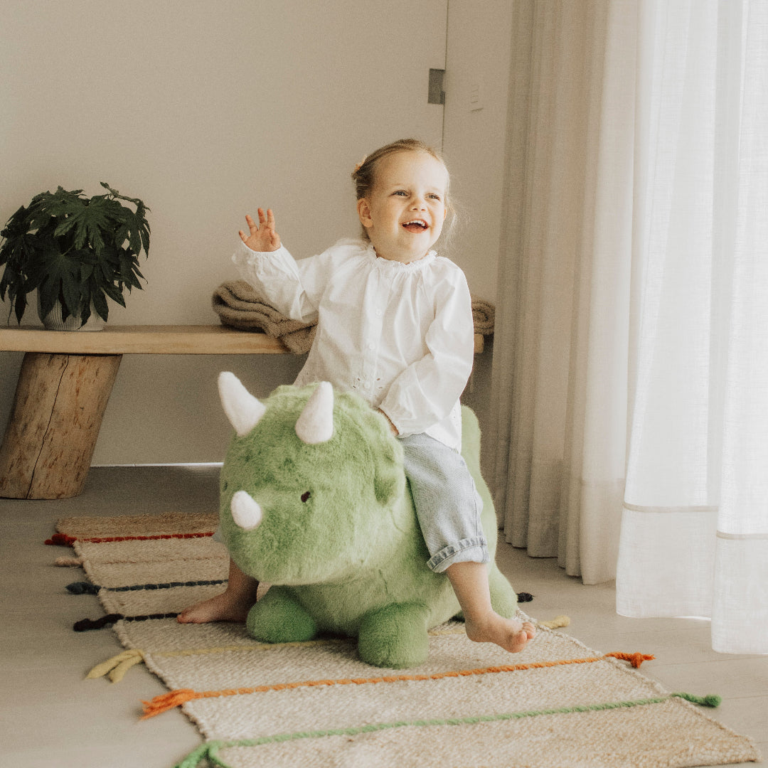 Een lachend meisje zit op het Wild & Soft groene pluche skippydier in de vorm van een dinosaurus en zwaait.