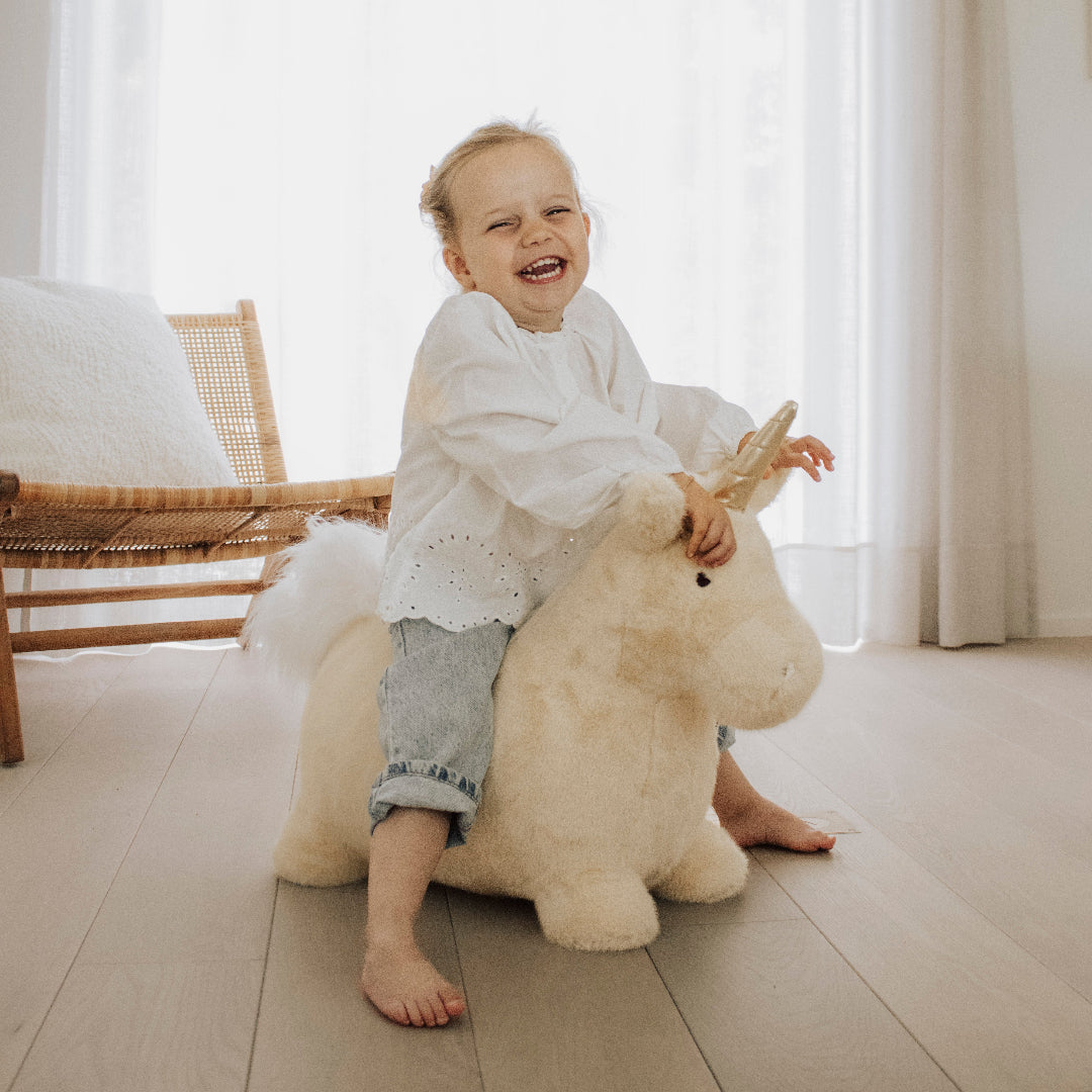 Een vrolijk peutermeisje in een wit shirt zit op het Wild & Soft beige pluche eenhoorn skippydier.
