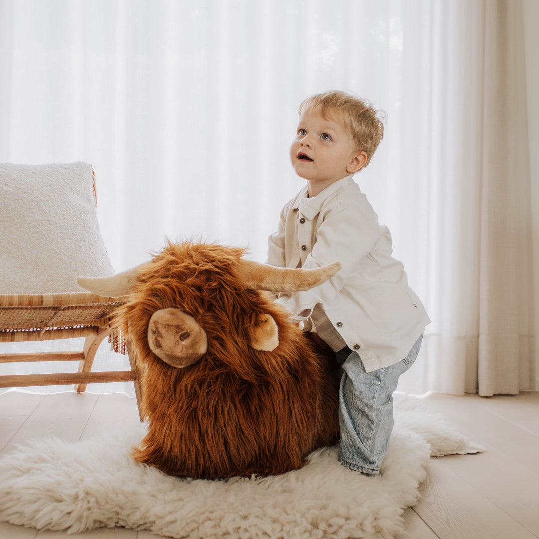 Een jong kind staat naast de Wild & Soft skippydier Schotse Hooglander op een wit kleed en kijkt omhoog.