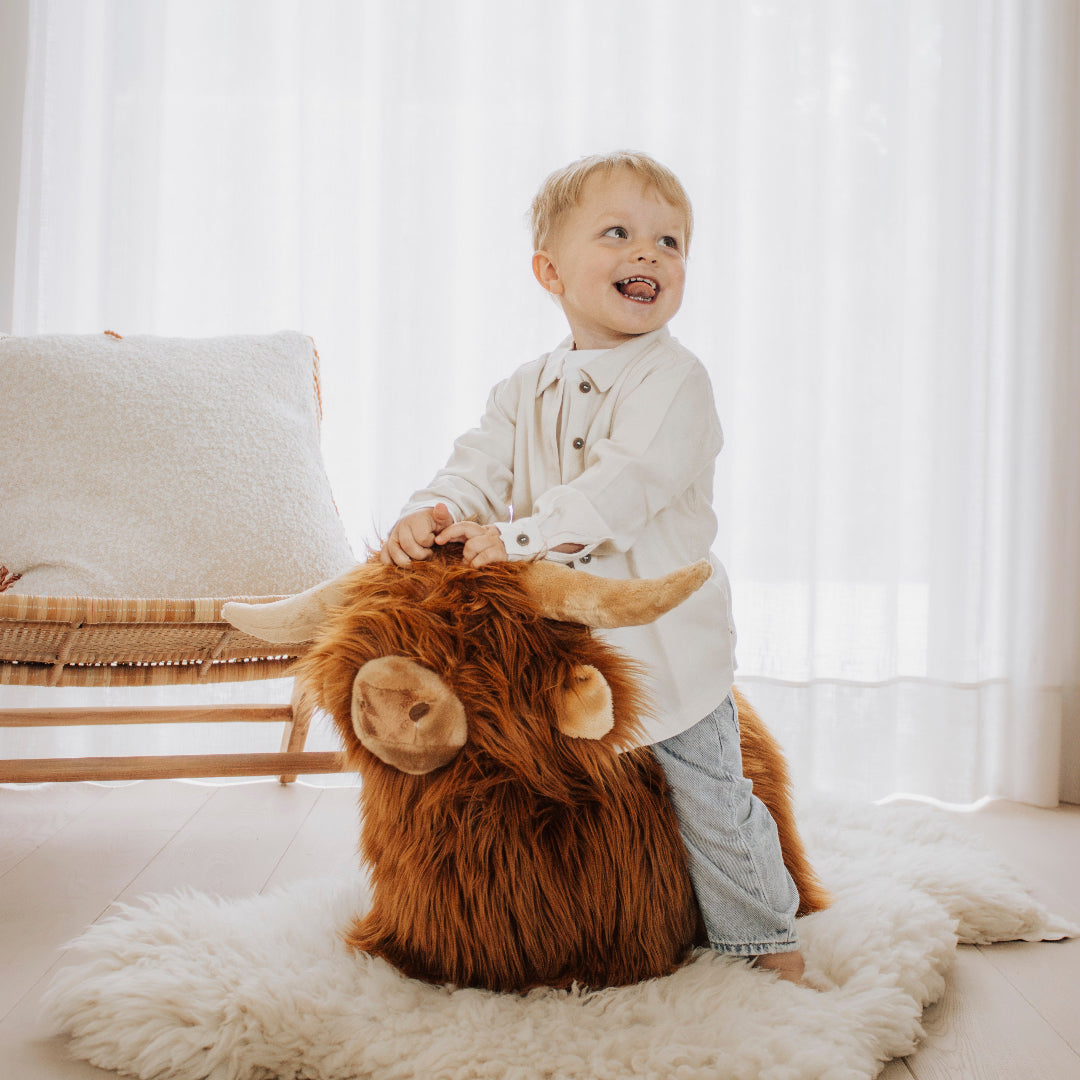Een lachend kind staat naast de Wild & Soft skippydier Schotse Hooglander op een wit kleed en houdt het vast.