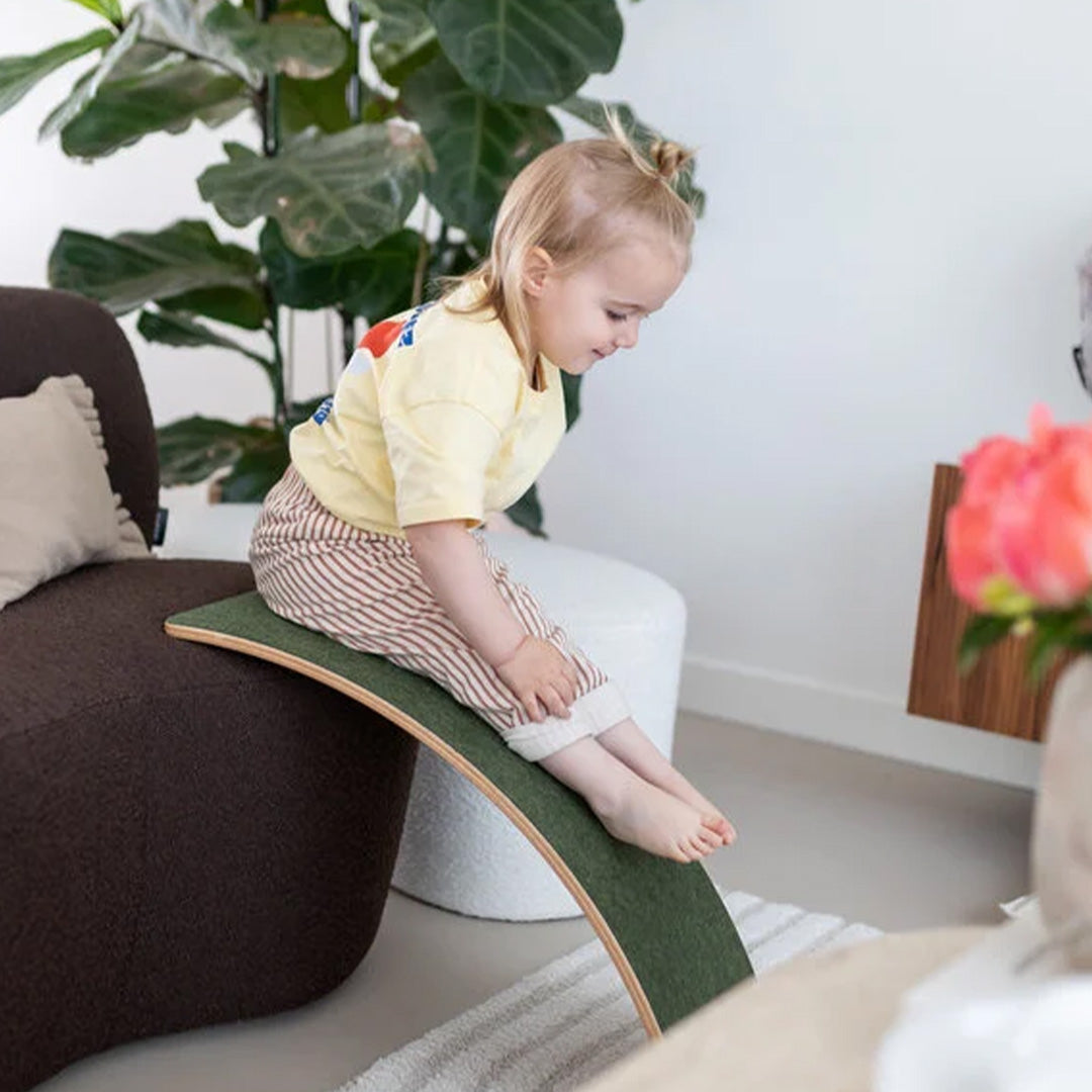 Een kind glijdt van een groen Wobbel balance board dat tegen een bank leunt.