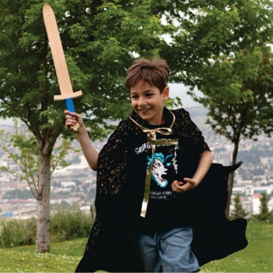 Een lachende jongen in een zwarte cape zwaait met het Woodsy houten speelgoedzwaard in een park.