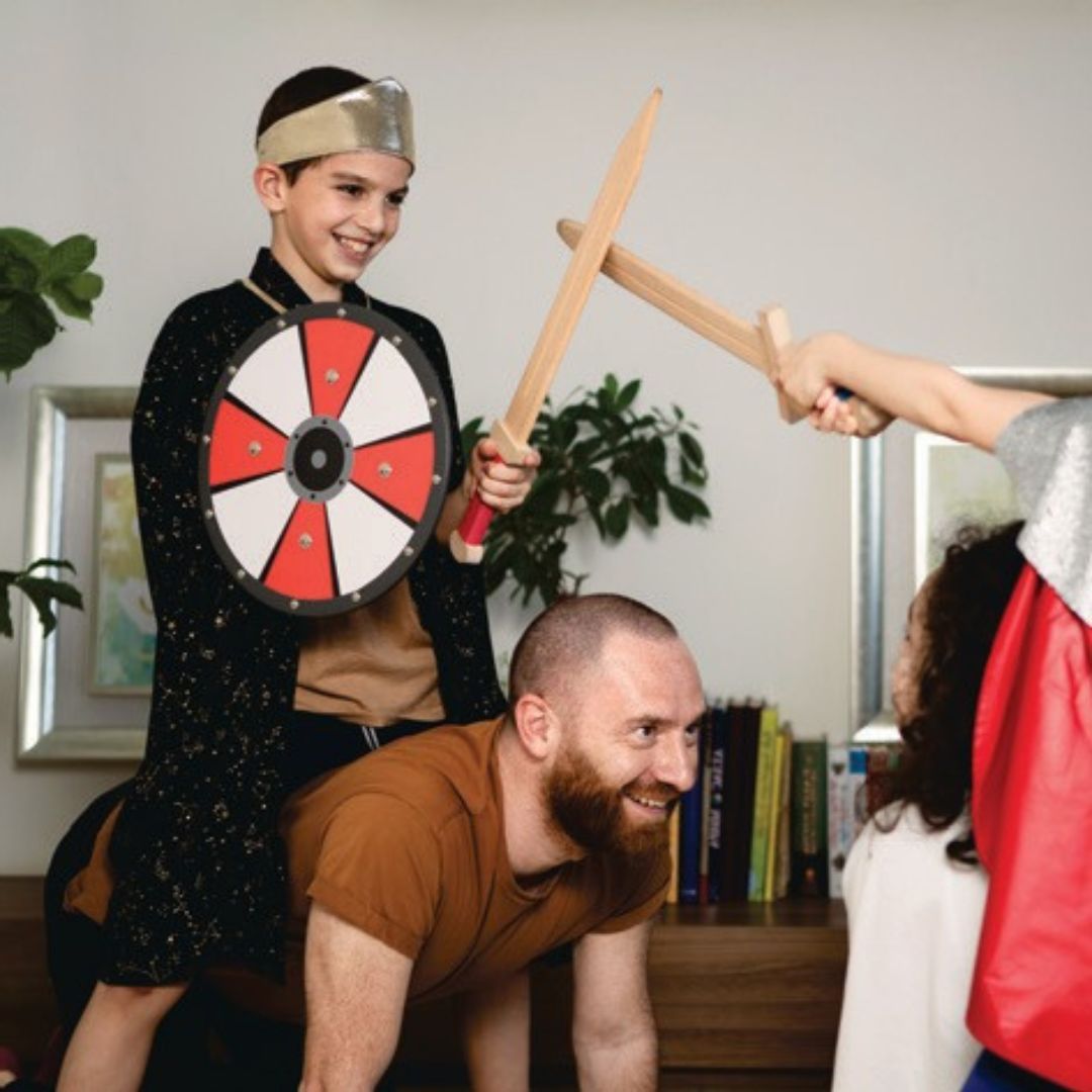 Twee kinderen en een volwassene spelen ridder; een jongen met kroon en schild zit op de rug van een man en houdt een houten zwaard vast.