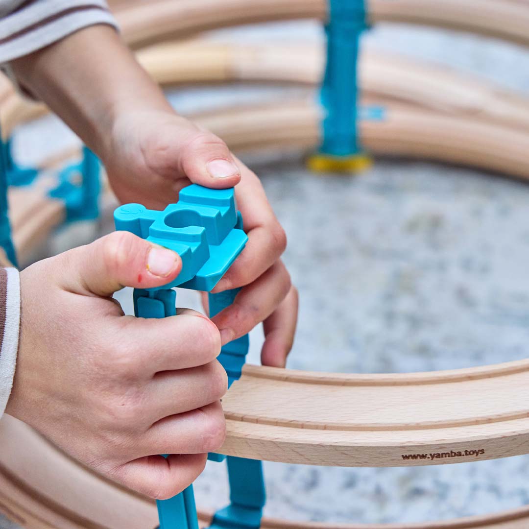 Kinderhanden die een blauwe Yamba Toys adapter verbinden met een houten treinbaanonderdeel.