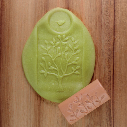 Een groene kleiafdruk van een boom en een beige Yellow Door kleistempel met een bladpatroon liggen op een houten ondergrond.