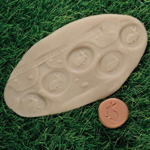 Een beige kleiafdruk met konijnen en een beige Yellow Door kleistempel met een konijnpatroon liggen op groen gras.