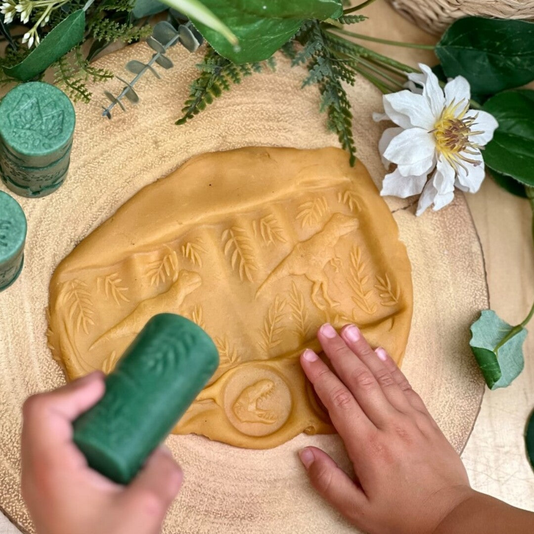 Kinderhanden gebruiken een groene Yellow Door kleistempel om dinosaurusafdrukken te maken in beige klei.