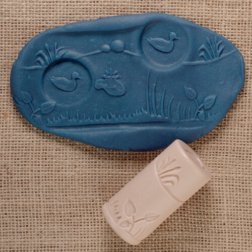 Een blauwe afdruk van vijverleven met eenden en riet, naast een beige kleistempel met rietpatroon.