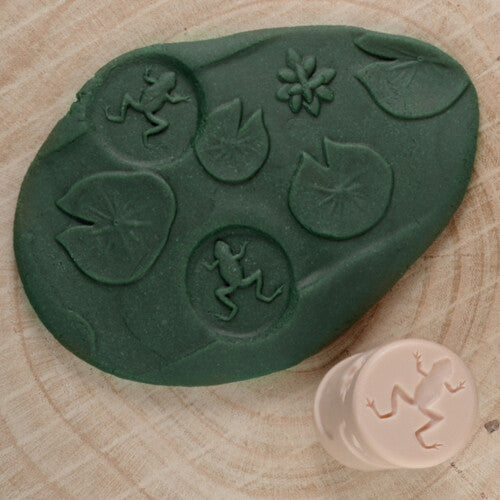 Een groene afdruk van vijverleven met kikkers en waterlelies, naast een beige kleistempel met kikkerpatroon.