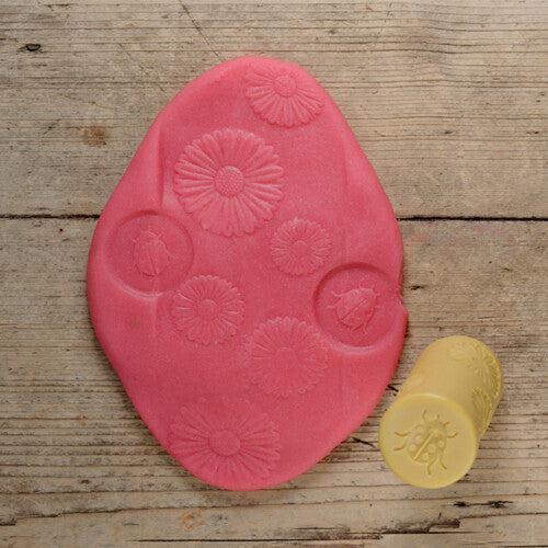 Een gele kleistempel met lieveheersbeestje ligt naast roze klei met afdrukken van bloemen en lieveheersbeestjes.