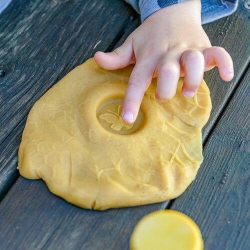 Een kinderhand drukt op gele klei met een vlinderafdruk, naast een gele kleistempel.