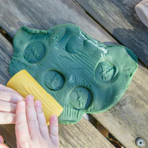 Kinderhanden rollen een gele kleistempel met graspatroon over groene klei.