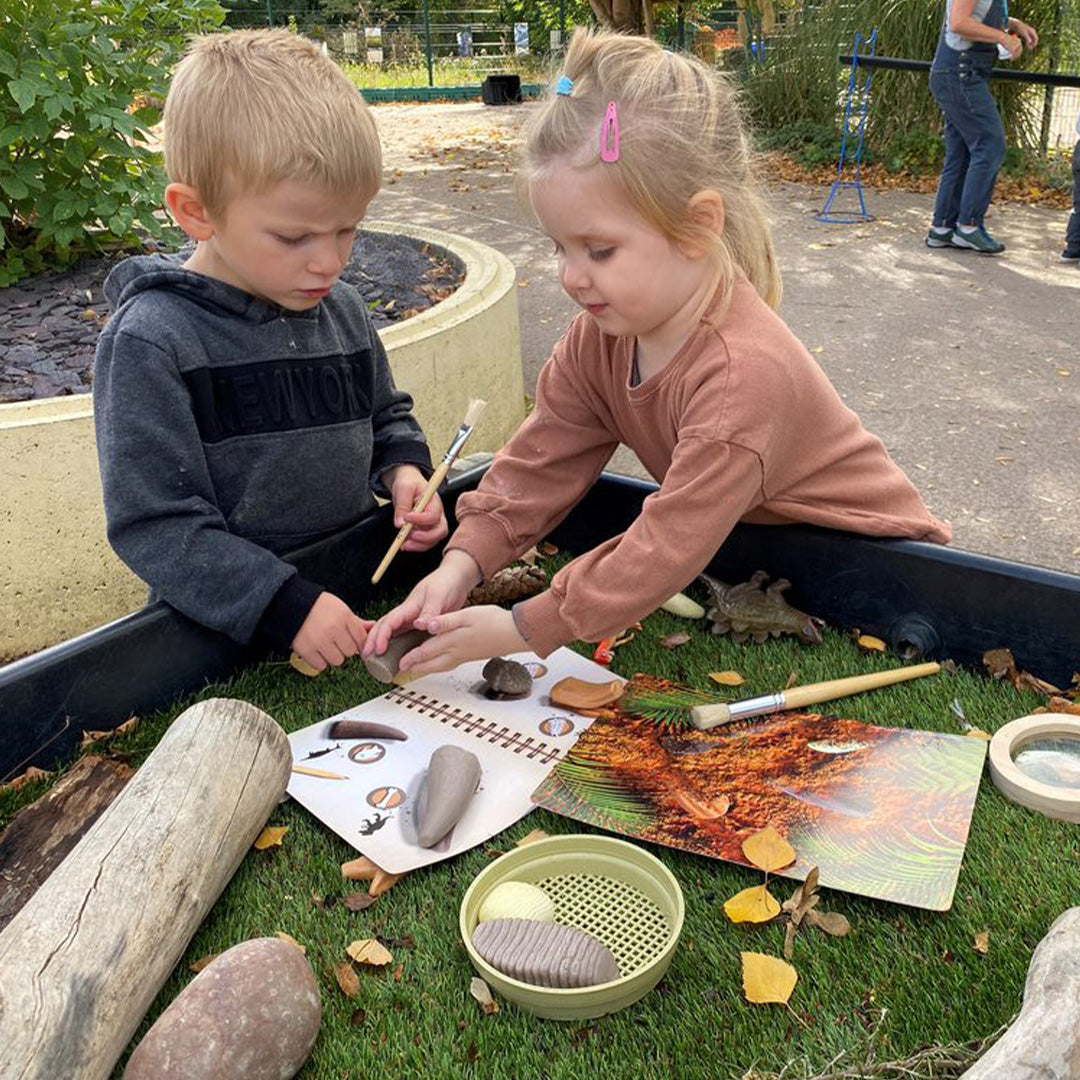Twee jonge kinderen spelen met de prehistorische tanden discovery set in een speelbak met kunstgras en bladeren.