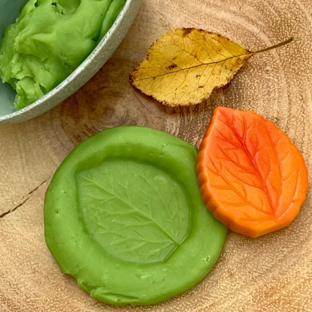 Een groene afdruk van een blad in klei, een oranje sensorische speelsteen en een echt geel herfstblad op een houten ondergrond.