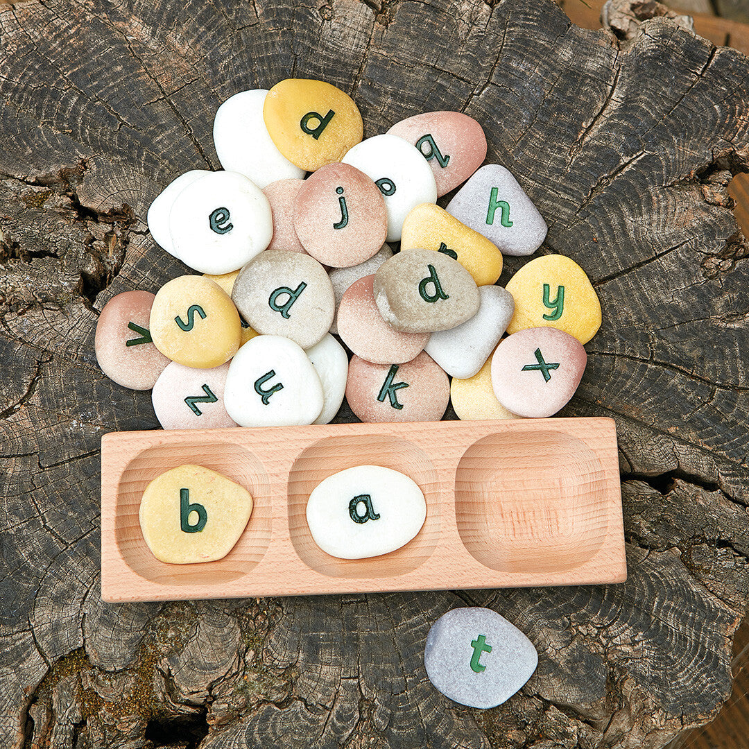 Houten sorteerbak met letterstenen die het woord 'bat' spellen, omringd door meer letterstenen op een boomstronk.