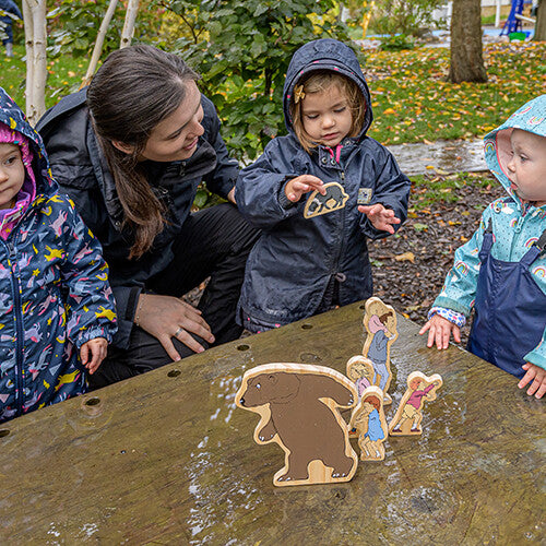 Een volwassene en drie kinderen spelen buiten met de Yellow Door houten 'We're Going on a Bear Hunt' speelfiguren op een houten tafel.