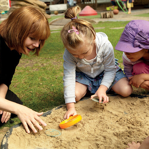 Een volwassene en twee kinderen spelen met de Yellow Door pootafdrukken speelstenen in een zandbak.