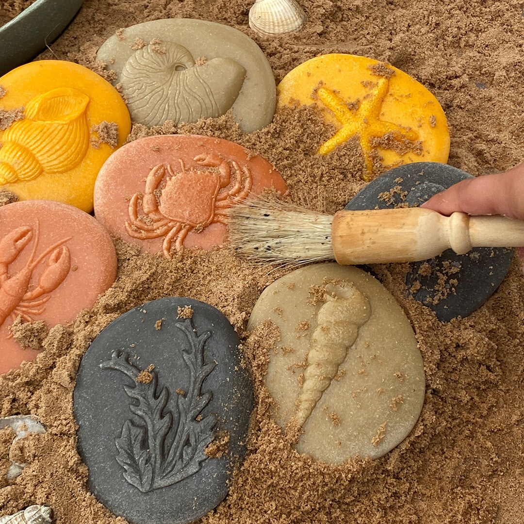 Een hand borstelt zand van een van de kustthema speelstenen met een klein kwastje.