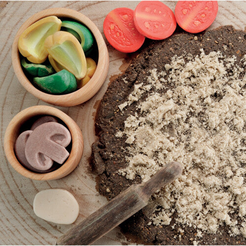 Een close-up van een modderpizza met zand, omringd door Yellow Door speelgoed pizza toppings van steen in houten kommetjes en een deegroller.
