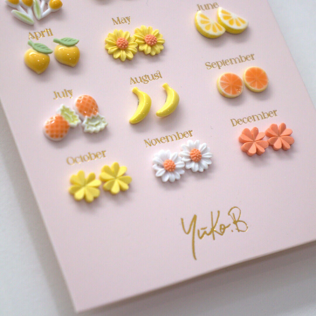 Detailopname van yuko b. kinder oorknopjes op een roze displaykaart, met fruit- en bloemendesigns voor de maanden juli tot en met december.