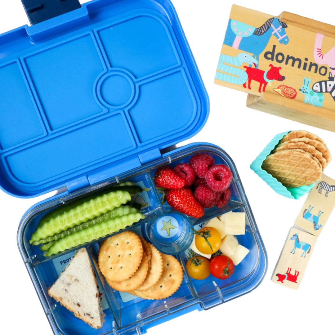 Open blauwe Yumbox broodtrommel voor kinderen gevuld met diverse etenswaren, met een dominospel ernaast.