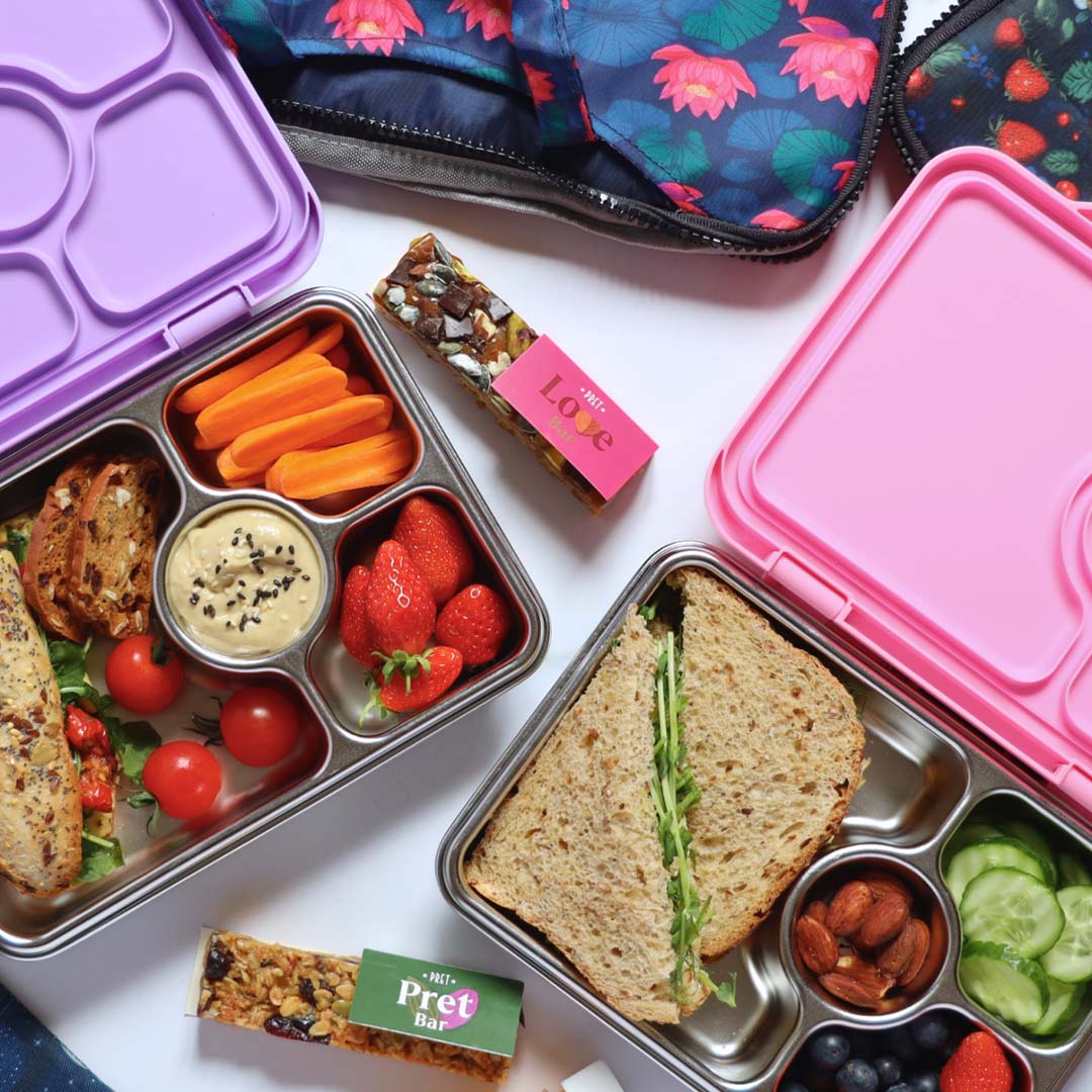 Twee open Yumbox bento broodtrommels, een paarse en een roze, gevuld met diverse etenswaren.