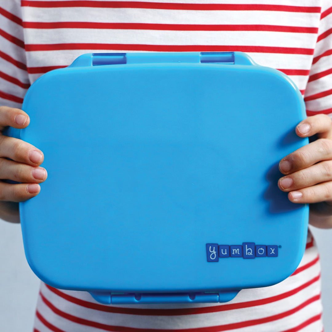 Een persoon houdt de gesloten blauwe Yumbox bento broodtrommel met logo vast.