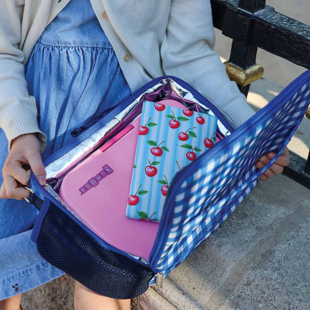 Een persoon houdt een open Yumbox lunchtas vast met daarin een roze lunchbox en een koelelement hoes met kersenpatroon.
