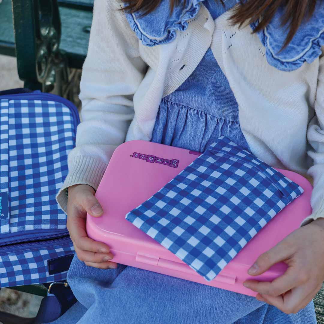 Een persoon houdt een roze Yumbox lunchbox vast met daarop een blauw-wit geruite koelelement hoes.