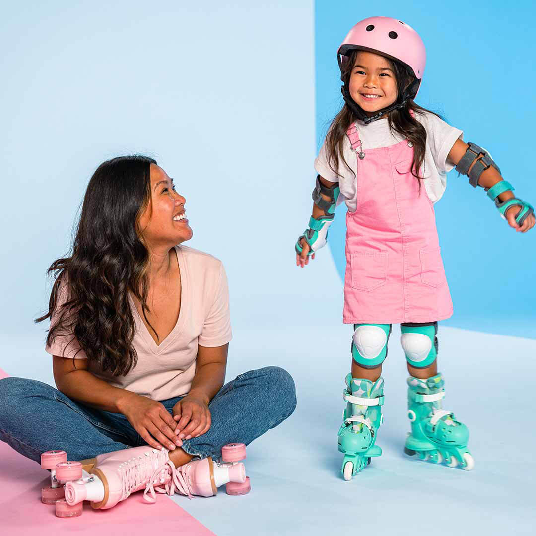 Een lachende vrouw kijkt naar een meisje met een roze helm en mintgroene kinder rolschaatsen.