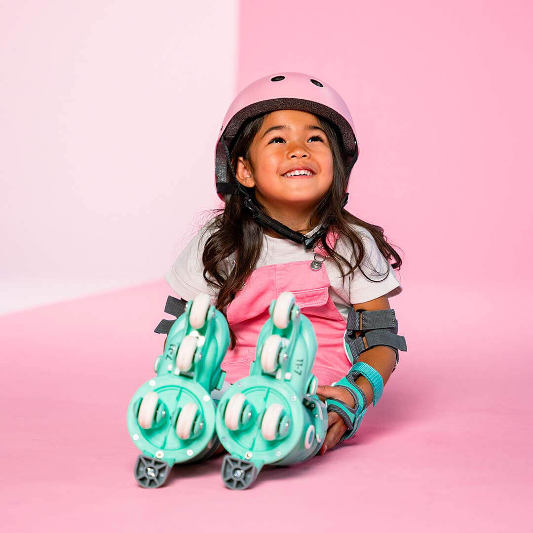 Een lachend meisje met een roze helm en beschermers zit naast haar mintgroene kinder rolschaatsen.