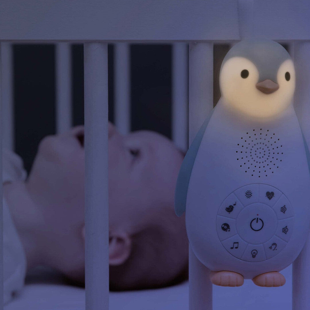 Het Zazu pinguïn nachtlampje hangt aan een wiegje en geeft zacht licht, met een slapende baby op de achtergrond.