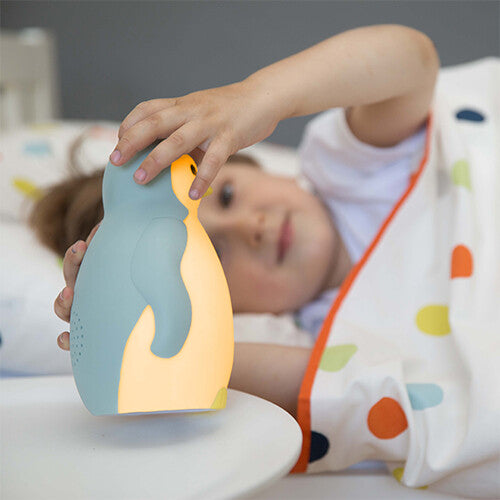 Een kind in bed raakt de Zazu slaaptrainer Pam pinguïn aan die geel licht geeft.