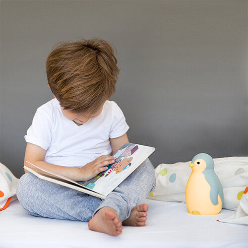 Een kind leest een boek op bed naast de Zazu slaaptrainer Pam pinguïn die geel licht geeft.