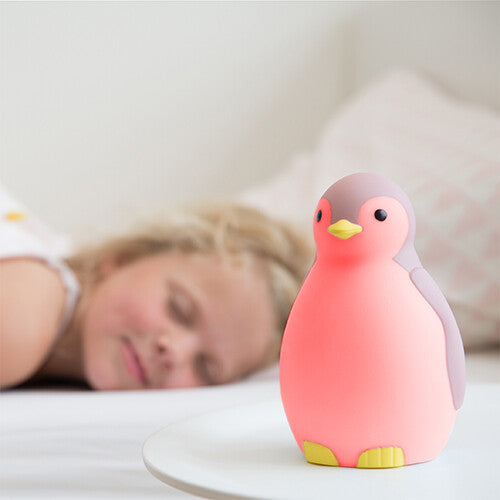 De roze Zazu pinguïn slaaptrainer geeft roze licht op een nachtkastje, met een slapend kind op de achtergrond.