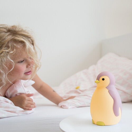 Een jong meisje kijkt vanuit bed naar de gele Zazu pinguïn slaaptrainer op een nachtkastje.