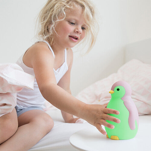 Een jong meisje in bed reikt naar de groene Zazu pinguïn slaaptrainer op een nachtkastje.