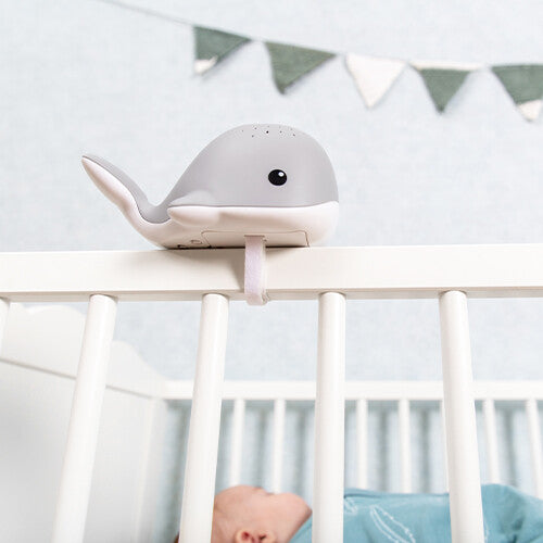 Zazu grijze walvis muziekdoos en lichtprojector bevestigd aan een babybedje, met een slapende baby erin.