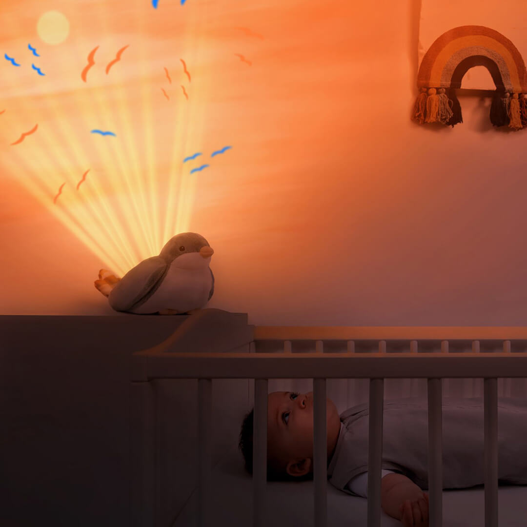Een baby kijkt vanuit de wieg naar de Zazu vogel muziekdoos die een zonsondergang en vogels op de muur projecteert.