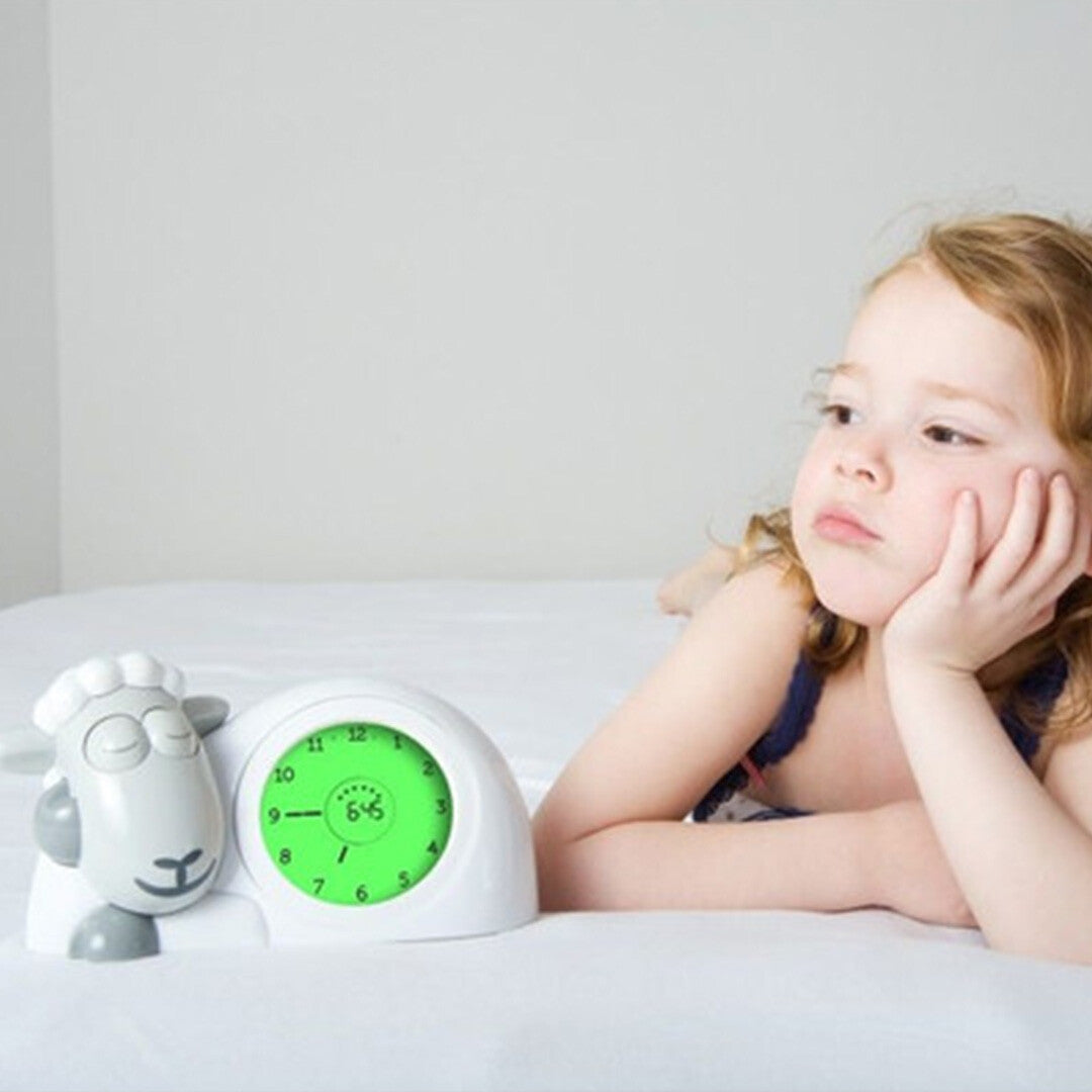 Een meisje ligt in bed naast de Zazu slaaptrainer en wekker Sam, die 6:45 aangeeft en gesloten ogen heeft.