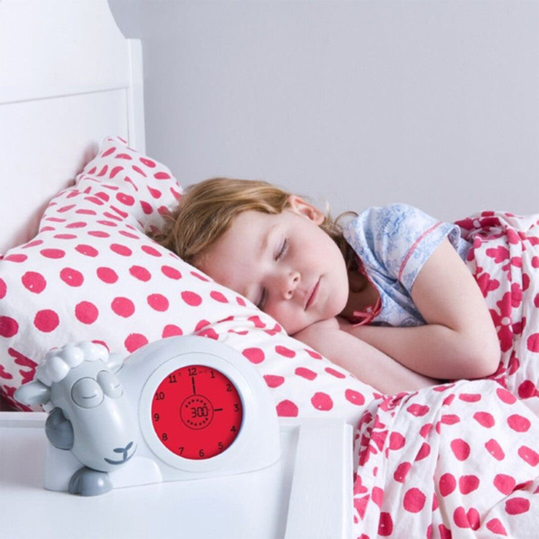 Een slapend meisje in bed naast de Zazu slaaptrainer en wekker Sam, die 3:00 aangeeft en gesloten ogen heeft.
