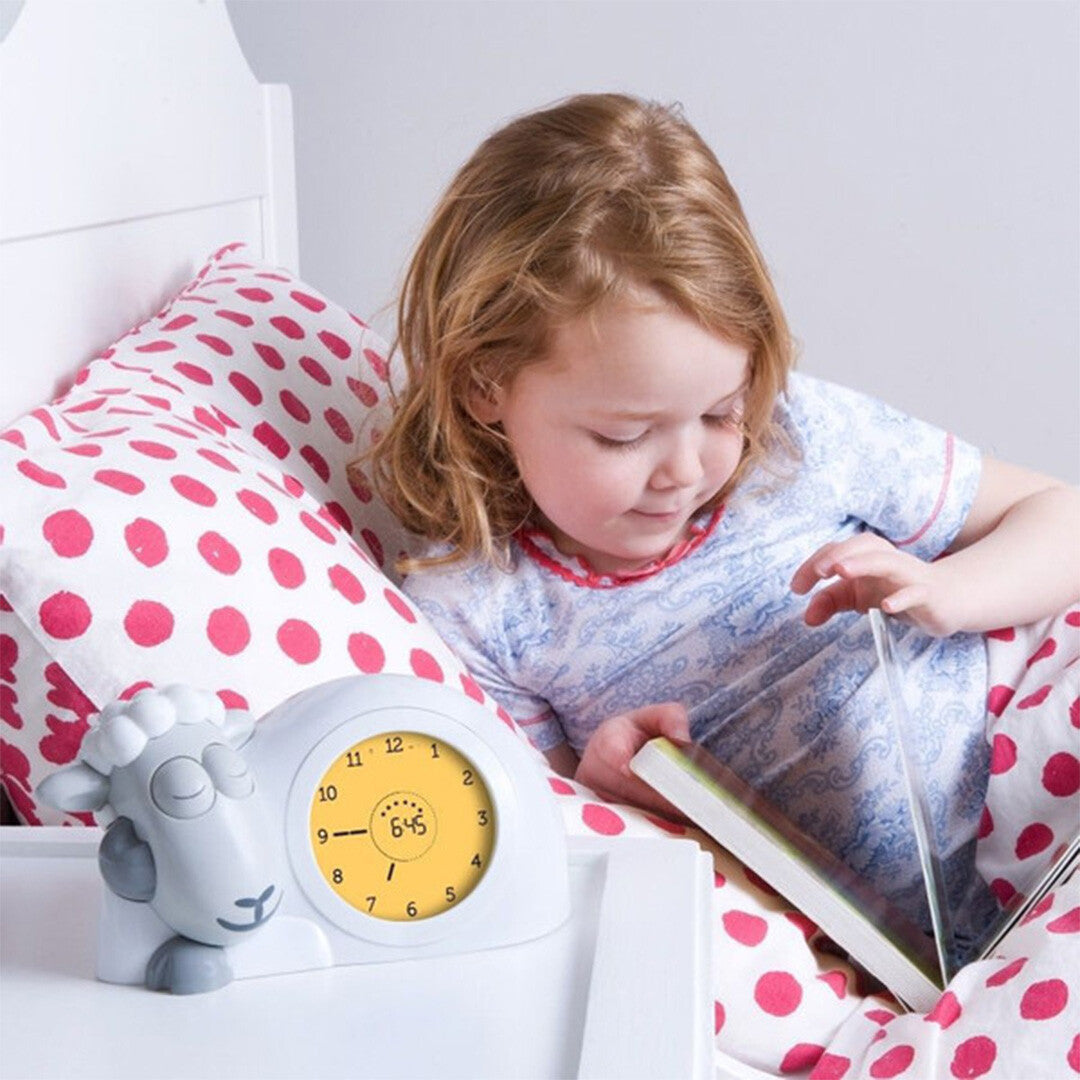 Een meisje leest een boek in bed naast de Zazu slaaptrainer en wekker Sam, die 6:45 aangeeft en gesloten ogen heeft.